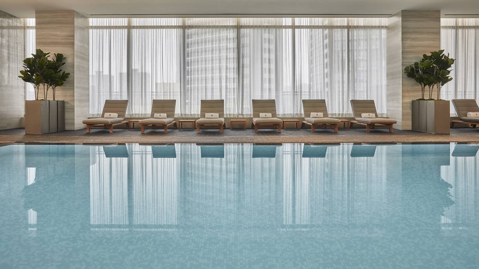 A wide angle view of the Four Seasons Toronto's indoor pool lined with cream lounge chairs and indoor potted trees. 