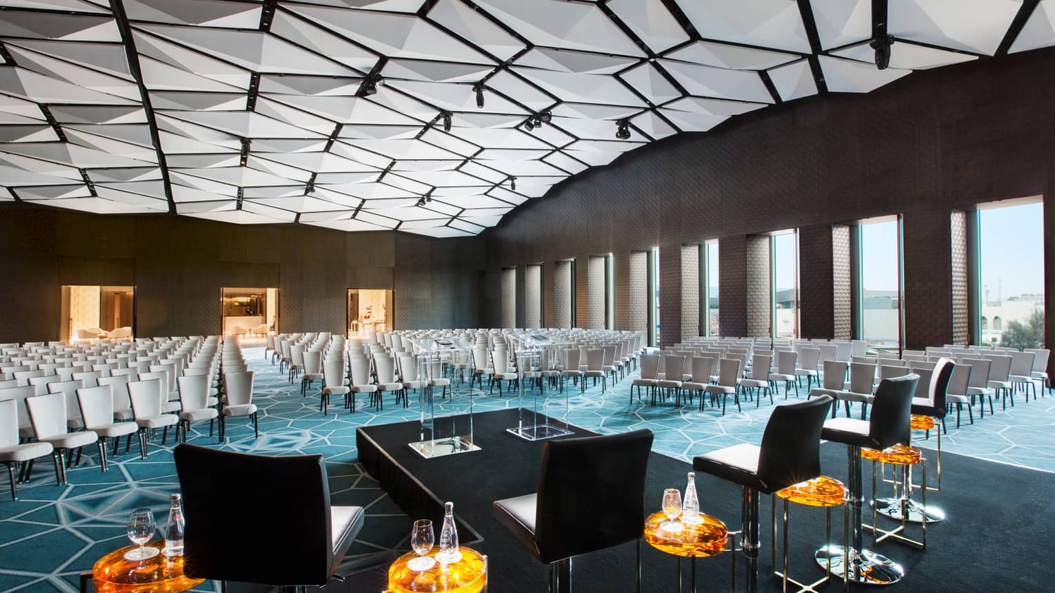 Rows of chairs fill large meeting room in front of stage, podium, geometric lights across ceiling