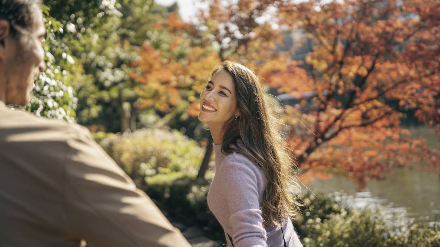 Smiling couple walks along the tree-lined pathway, pond and fall-hued trees in background