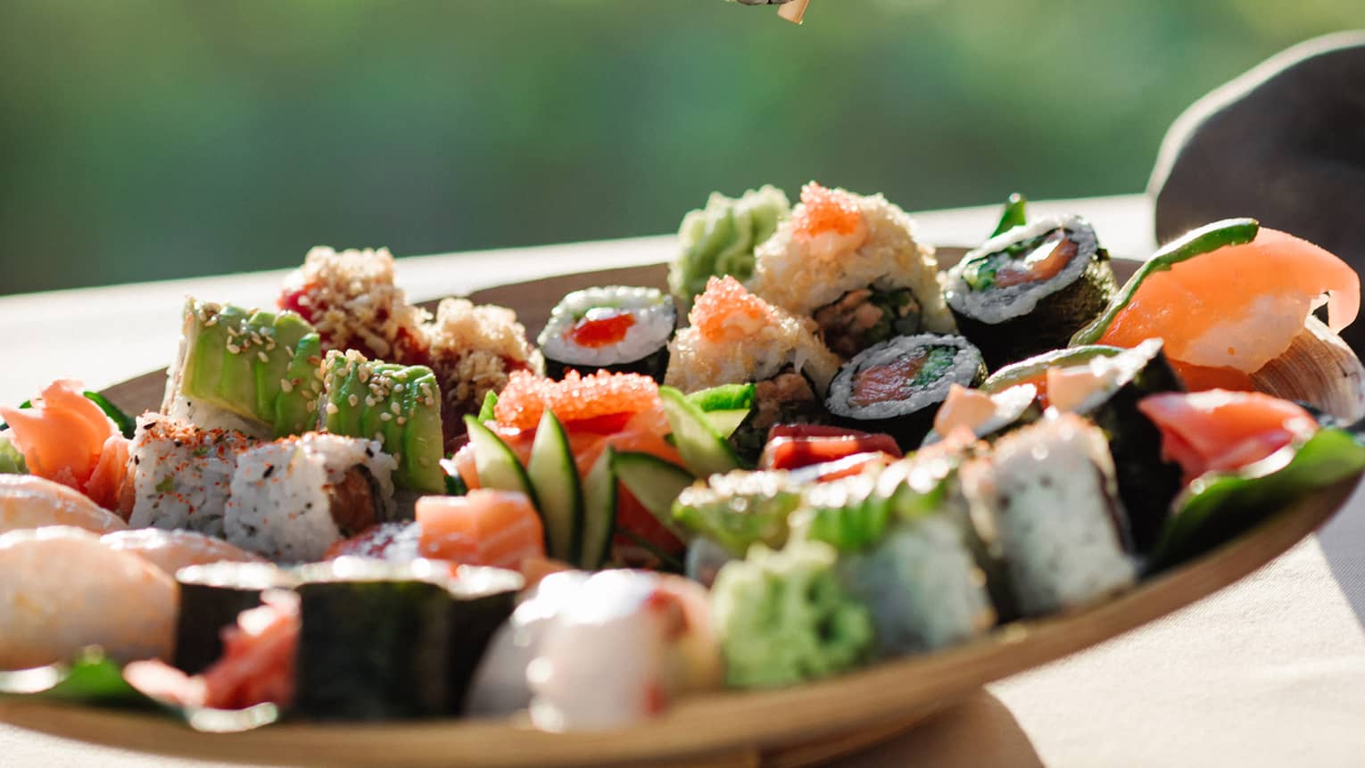Hand pinching chopsticks, holding a piece of sushi over a plate of assorted sushi