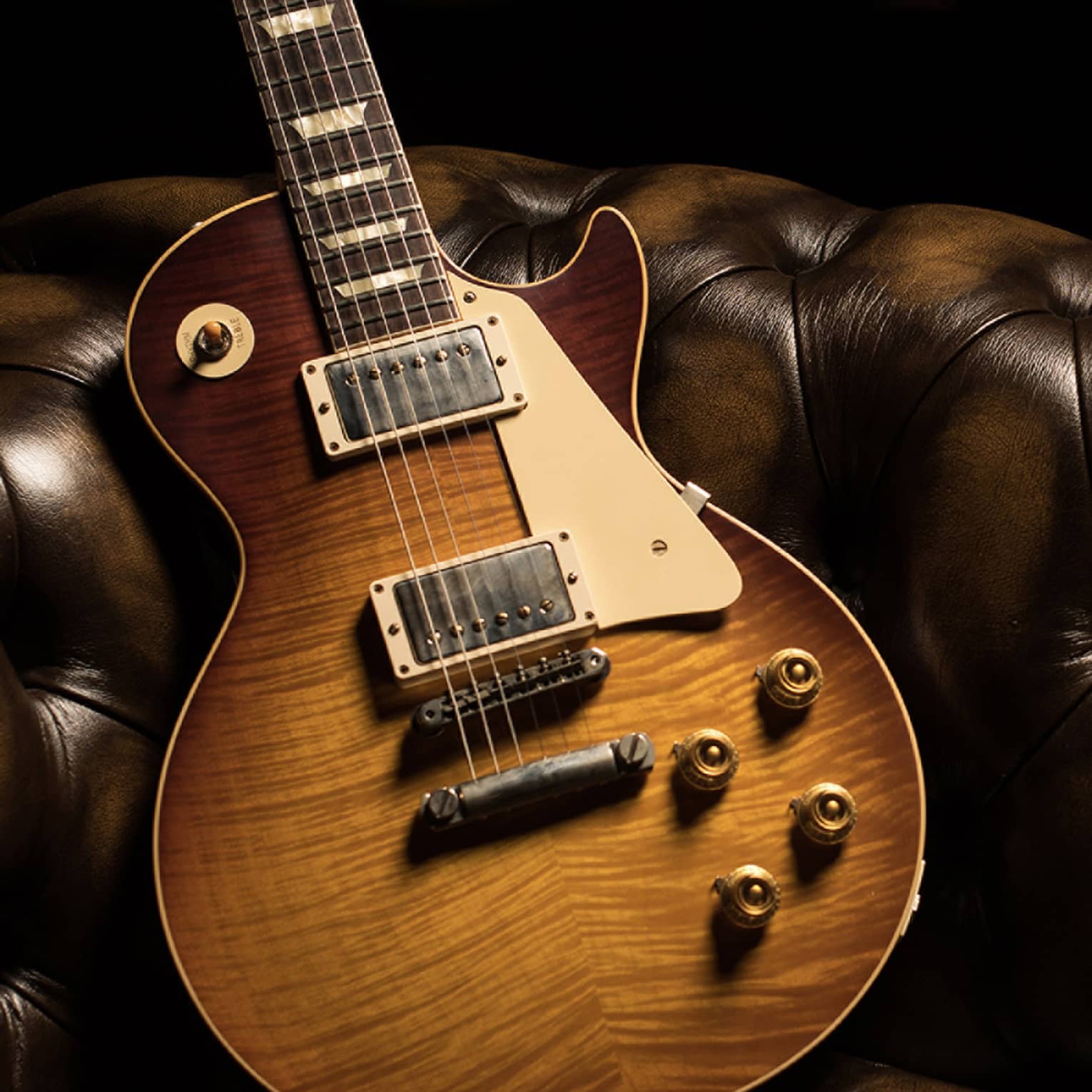 Close-up of a sunburst-finished electric guitar, its glistening body leaning diagonally against a brown leather tufted couch.