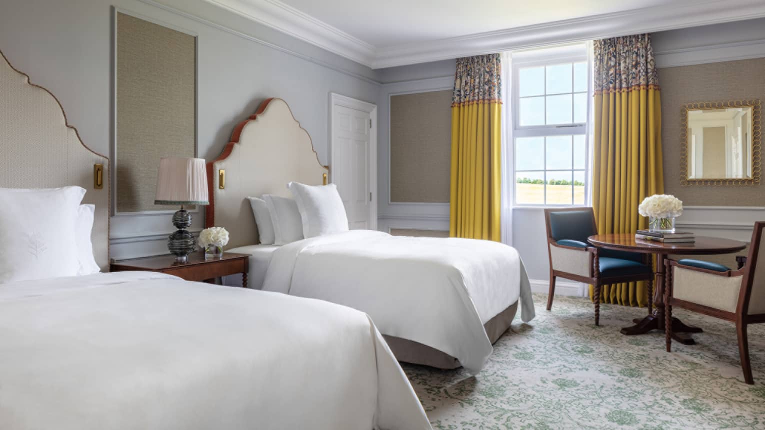 Guest room with two Double beds and twin tufted headboard, small table and two chairs, paisley carpeting, window with yellow drapes, mirror on wall