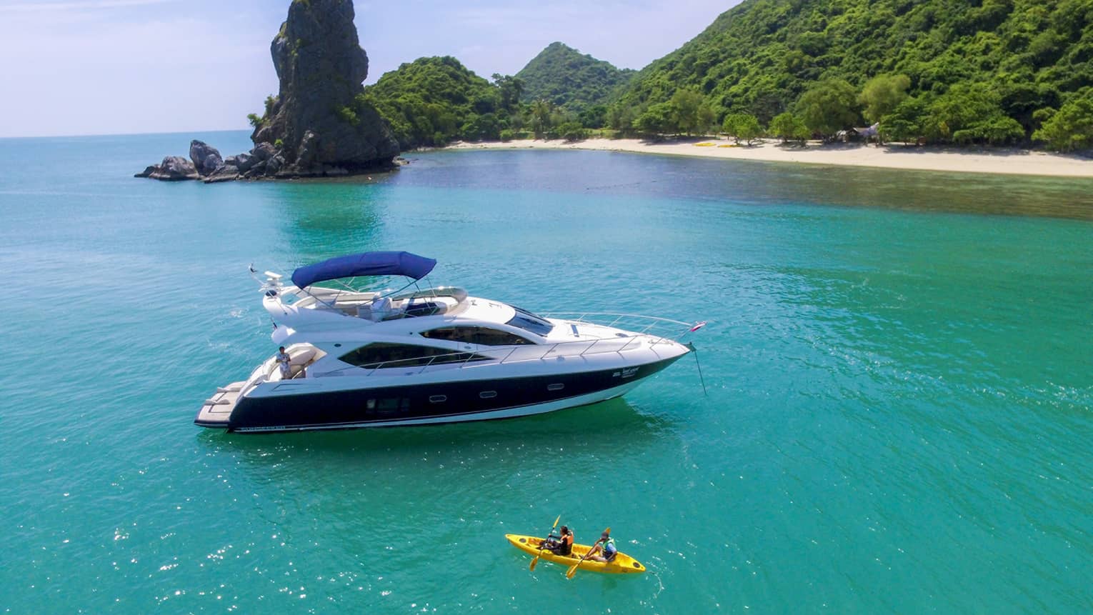 Two people in yellow kayak in front of large white yacht by island in blue ocean