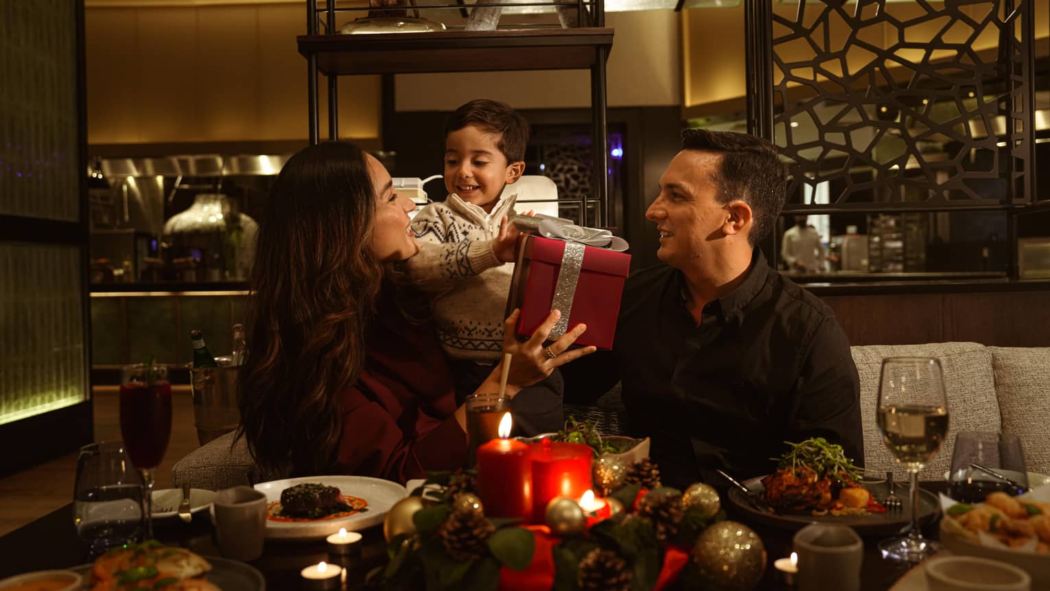 Two adults and a young child sit at a festive dining table opening presents
