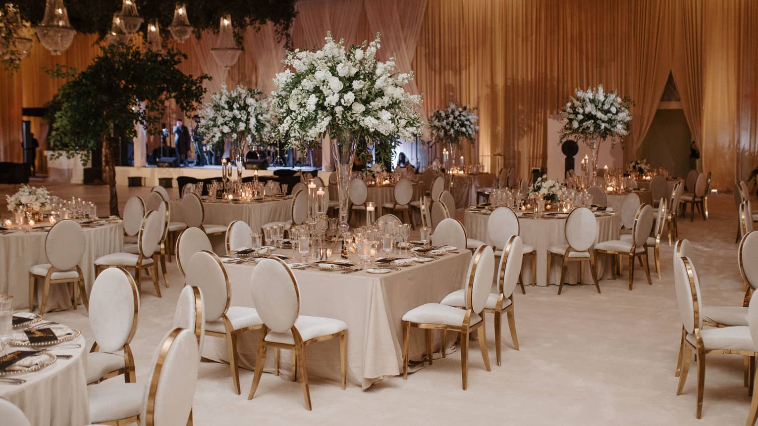 Ballroom decorated for a wedding reception with white floors, white-cloth tables and white dining chairs, with large white-and-green floral centrepieces at each table