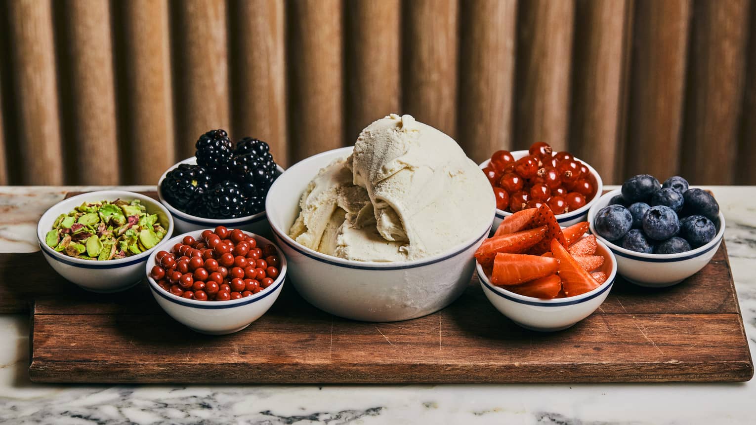 Large bowl of ice cream in centre of cutting board with two bowls of berries and one of nuts on left and three bowls with berries on right