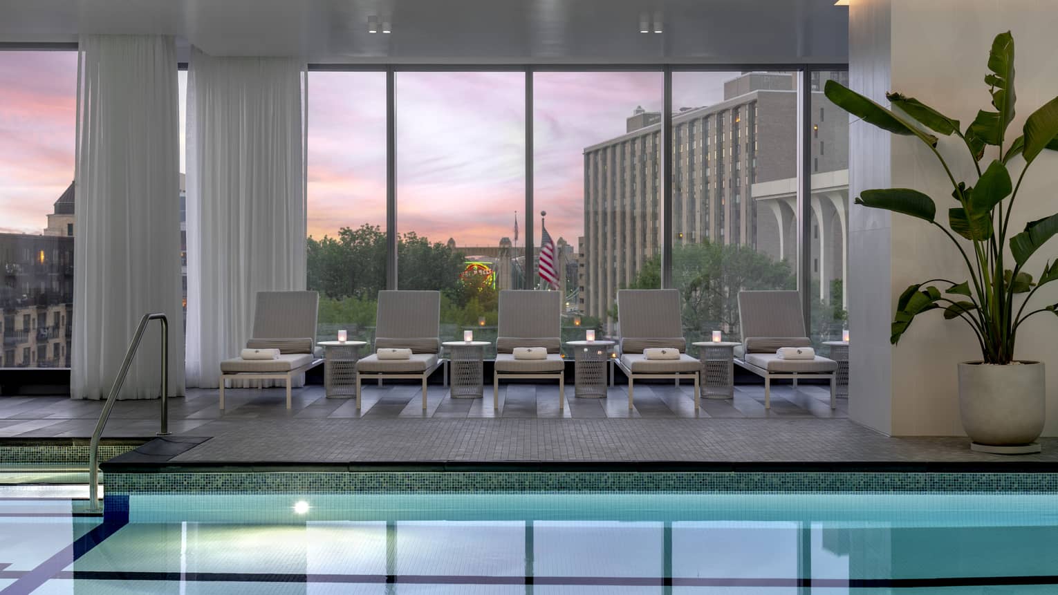 An indoor pool with dim lighting, plants, and lounge chairs with towels.