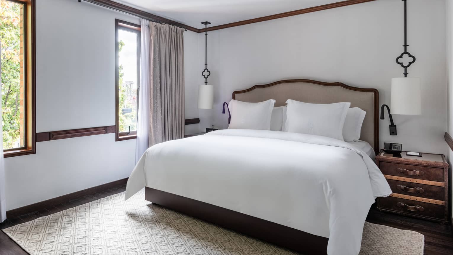 Guest room with king bed and white linens, tufted headboard, wooden side tables, handing lamps, area rug, twin windows
