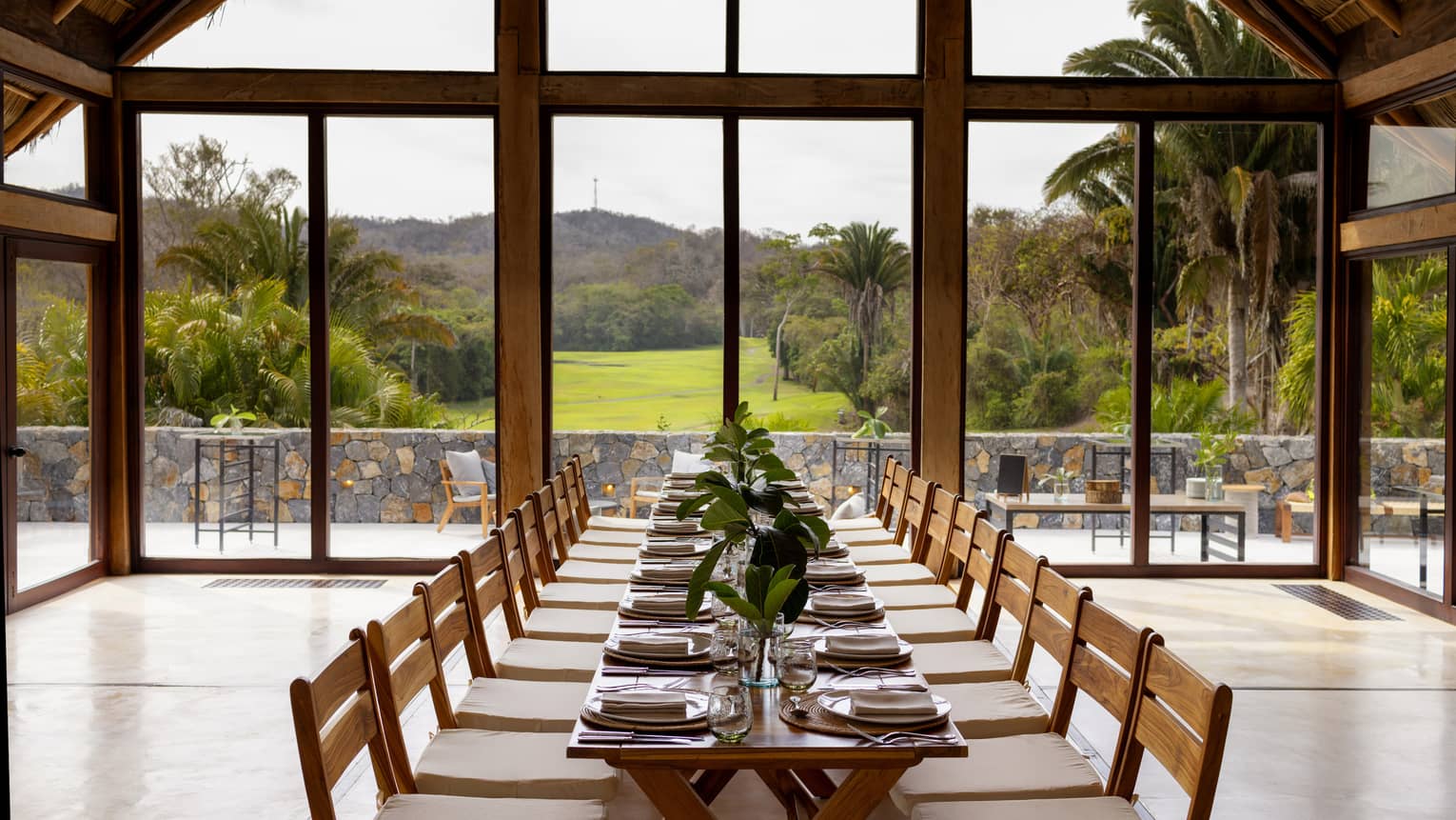 Room with floor-to-ceiling windows and a view of greenery and the mountains, with a rectangular table and place settings.