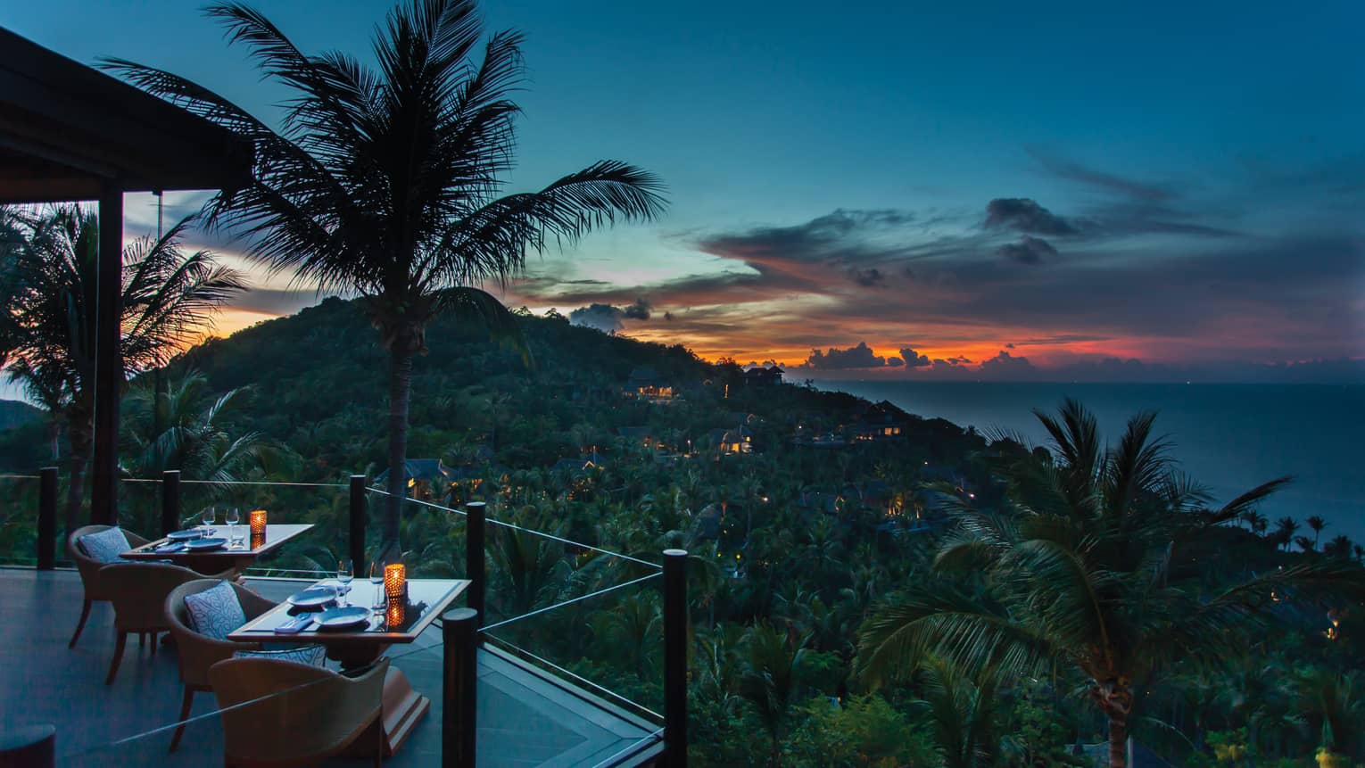 Corner of extended balcony with dining tables and chairs at dusk, expanse of forest, palm trees