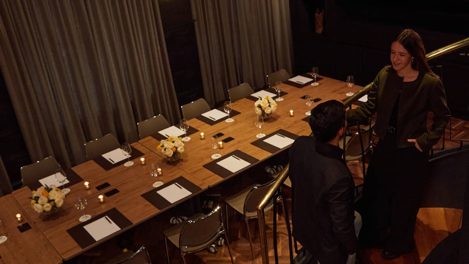 Two people talk on a curved stairwell with a long rectangular table set below them in a private meeting room
