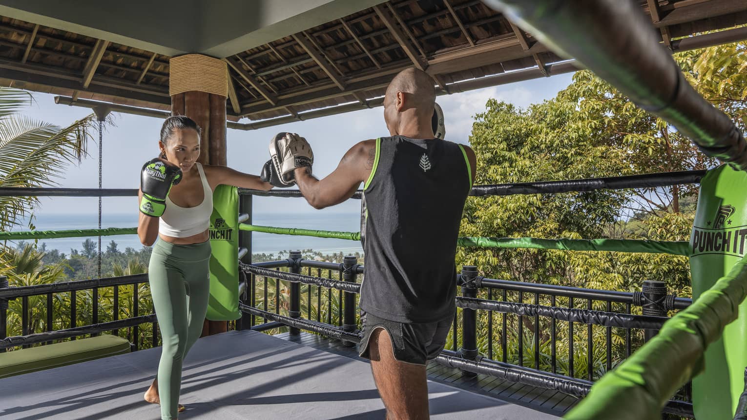 Two people practise Muay Thai in an outdoor boxing ring nestled in the treetops and overlooking the forest and the ocean.