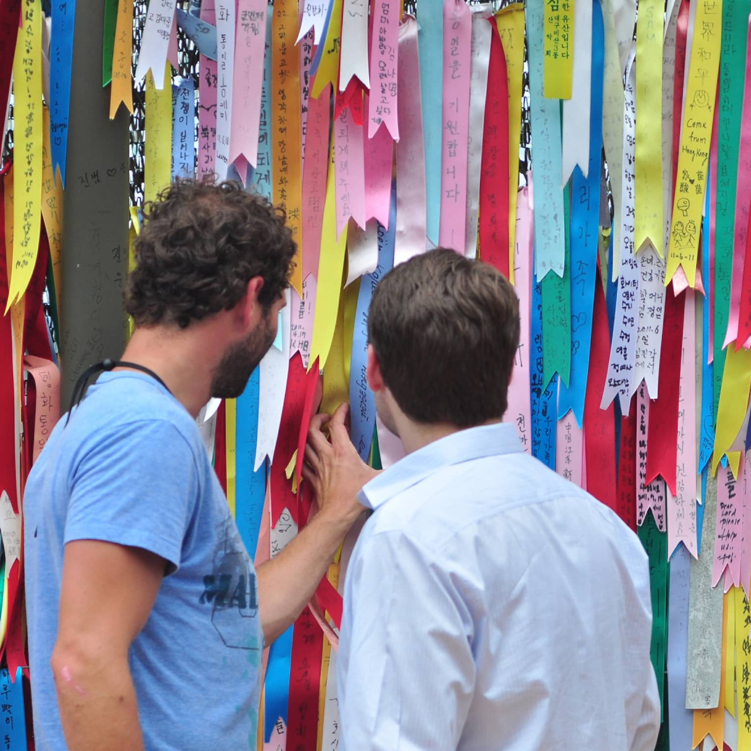 Two men looking at barbwire fence entirely covered with colourful ribbons with Korean text expressing wishes for world peace.