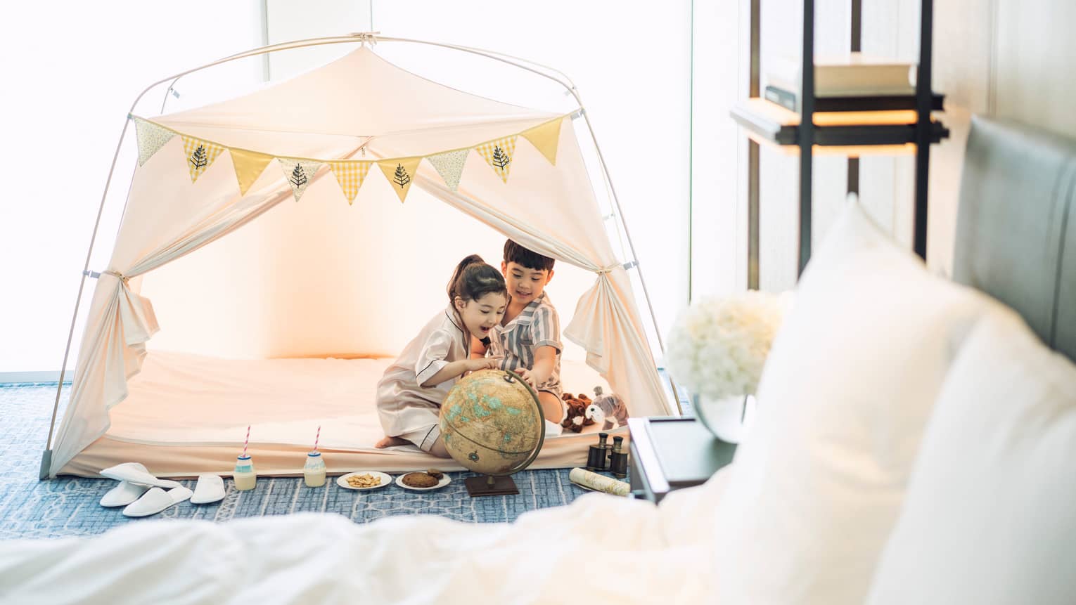 Two children wearing white bathrobes play in white tent in hotel room by bed