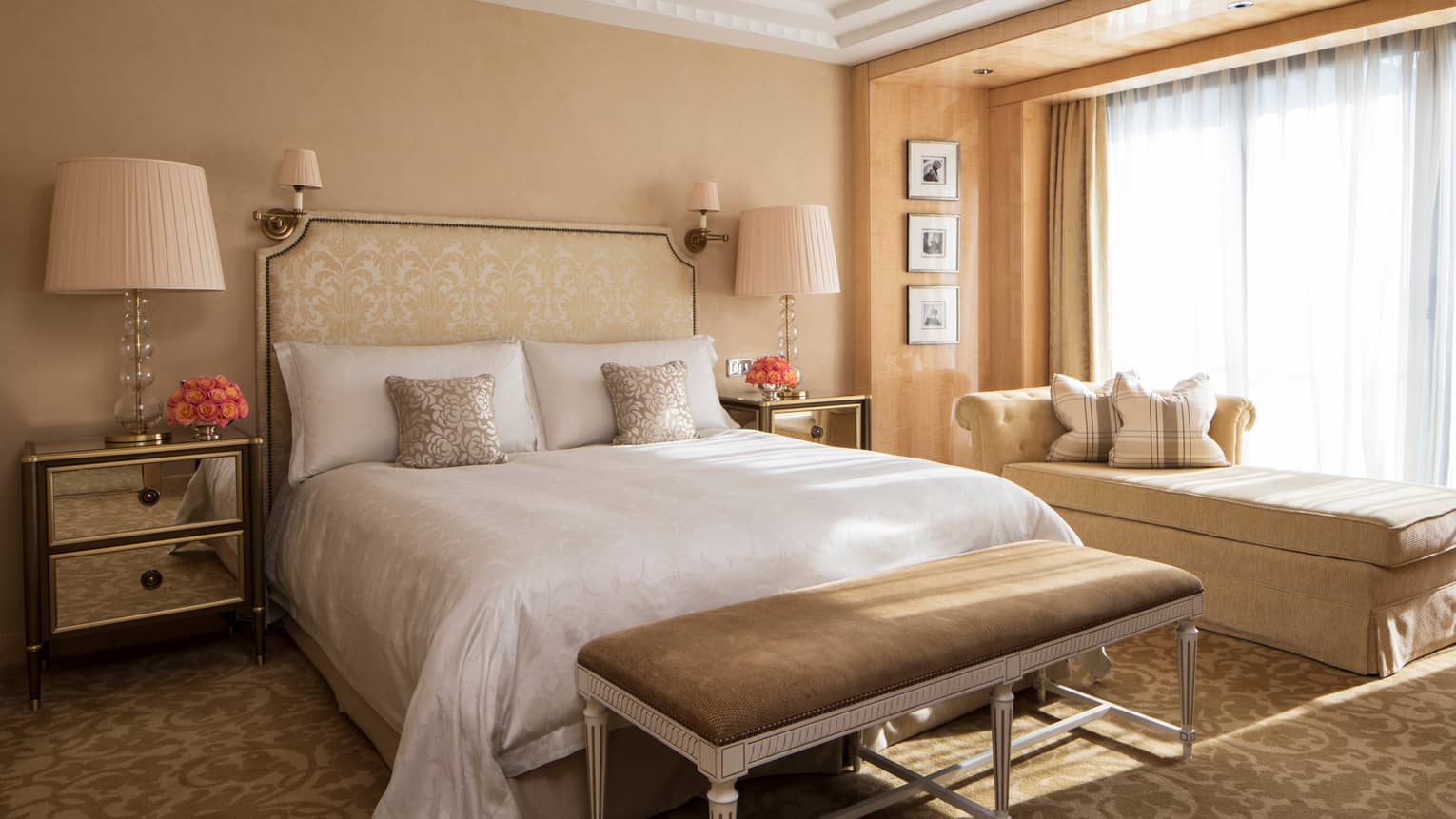 The Park Suite at Four Seasons London Park Lane in Mayfair offers a luxuriously appointed bedroom. It features a large bed with a decorative gold and cream headboard and elegant matching linens. Bedside tables with mirrored surfaces hold decorative lamps with soft pink shades, enhancing the room's warm and inviting ambiance. A long bench at the foot of the bed matches the golden upholstery of a nearby chaise lounge, which sits beside a window draped with sheer curtains, offering views of the serene outdoor setting. The room's refined decor is completed with a patterned carpet and tastefully framed black and white photographs on the wall.