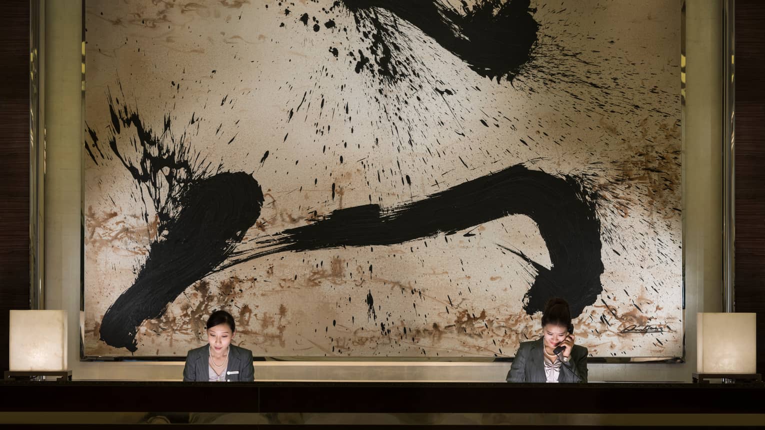 Two hotel reception staff at long desk under large black-and-white abstract painting