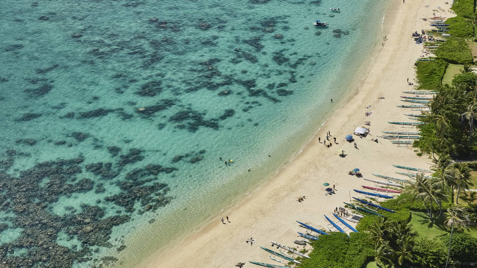 Aerial view of white sand beach lined with kayaks, blue lagoon with coral reef