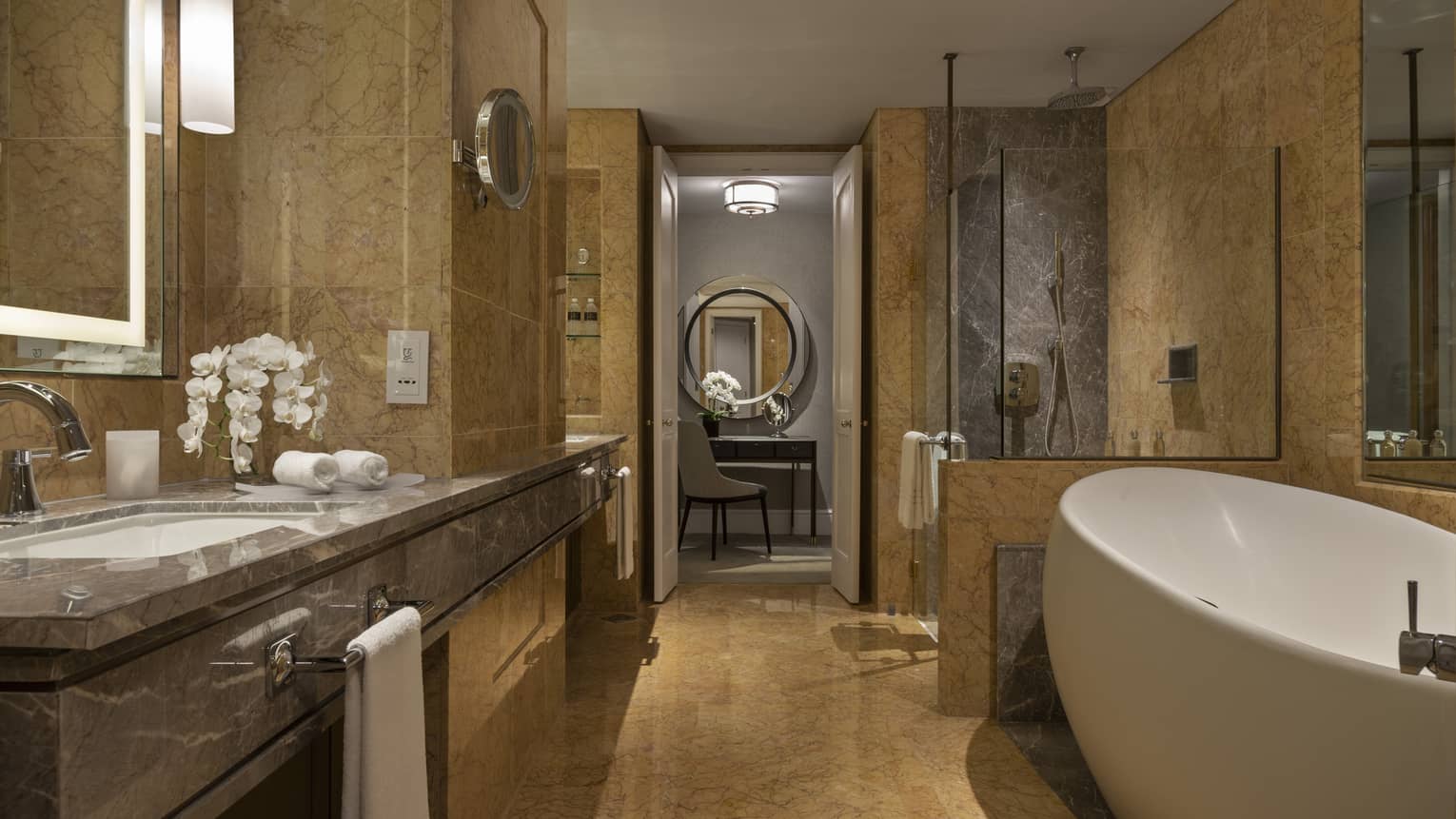 A marble bathroom with a freestanding bathtub, spacious vanity with double sinks and glass-enclosed shower. A vanity table with a circular mirror is visible through the open doorway.
