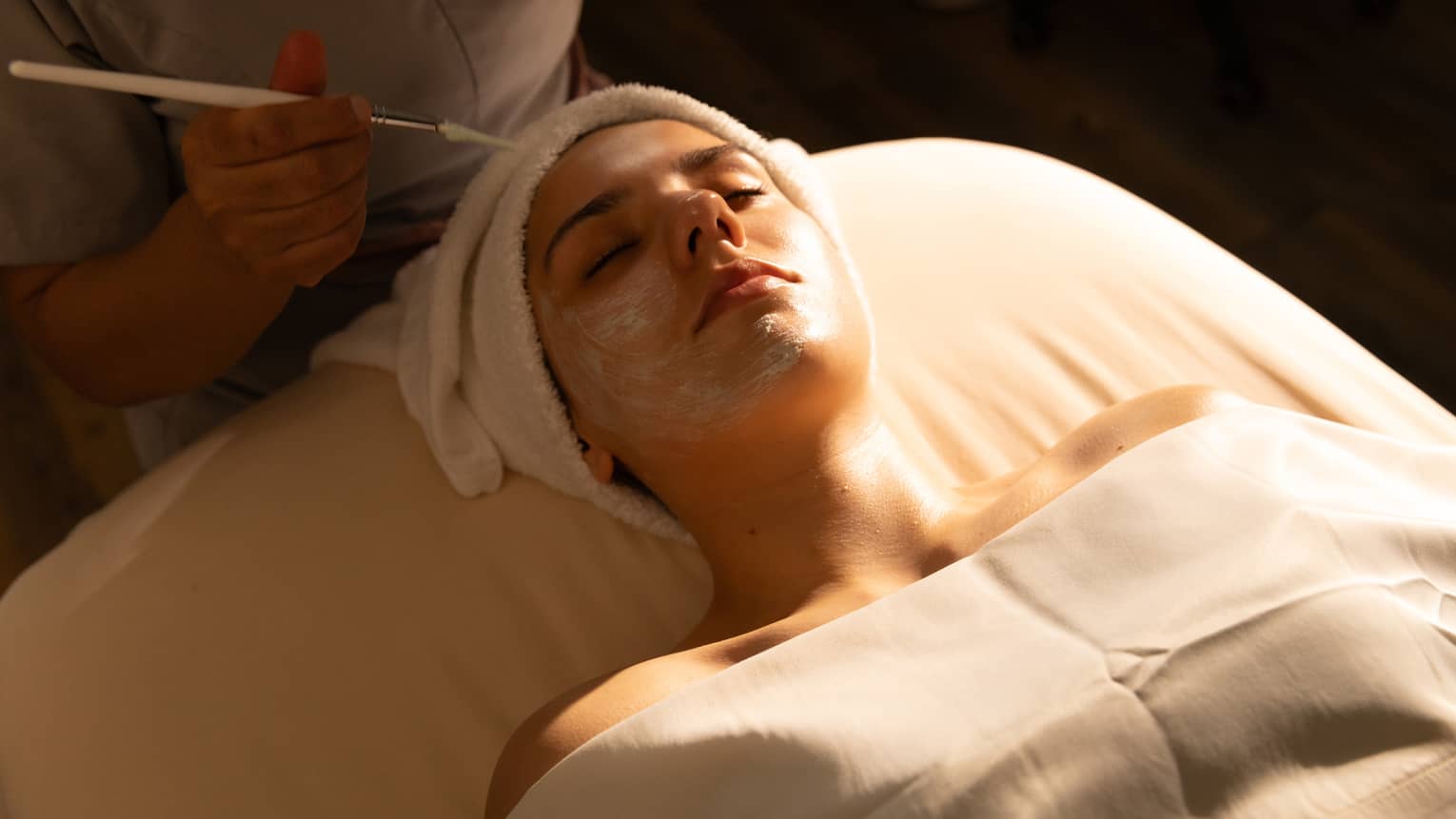 A person with draping over their body and a towel wrap on their head lies on a spa treatment table and receives a facial treatment.