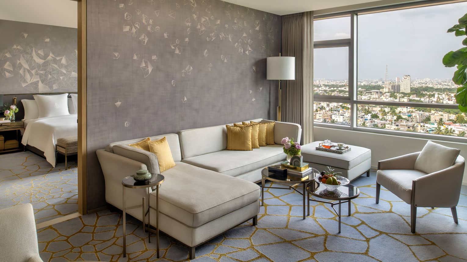Guest room living area with cream-coloured sectional sofa and matching armchair, window with city views, doorway to bedroom