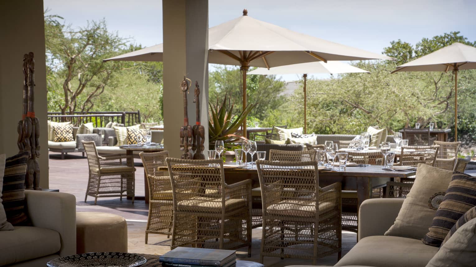 Wooden tables and chairs with large umbrellas, couches on outdoor patio with statues and decor