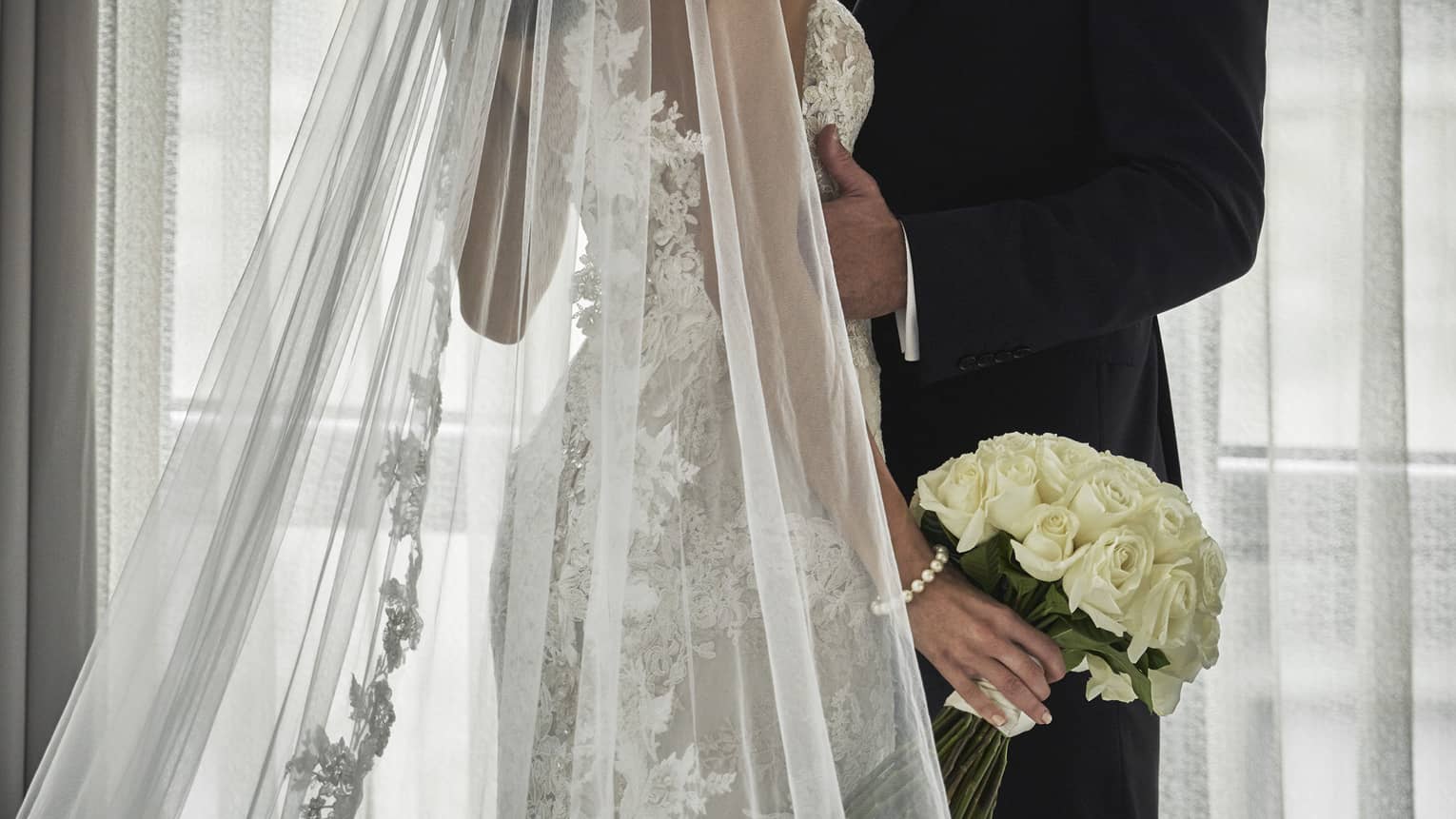 Groom and bride with long lace veil and white wedding bouquet embrace