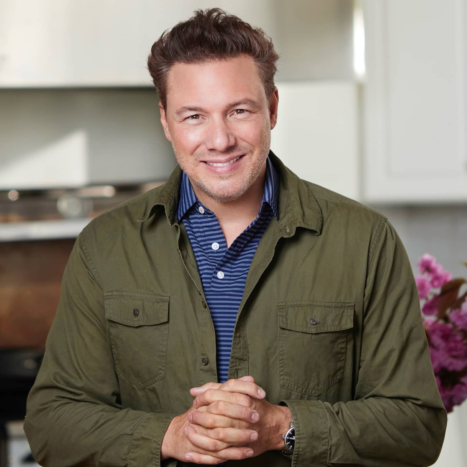 Rocco DiSpirito wearing a blue and white shirt with a green jacket and smiling with their hands clasped in front of them.