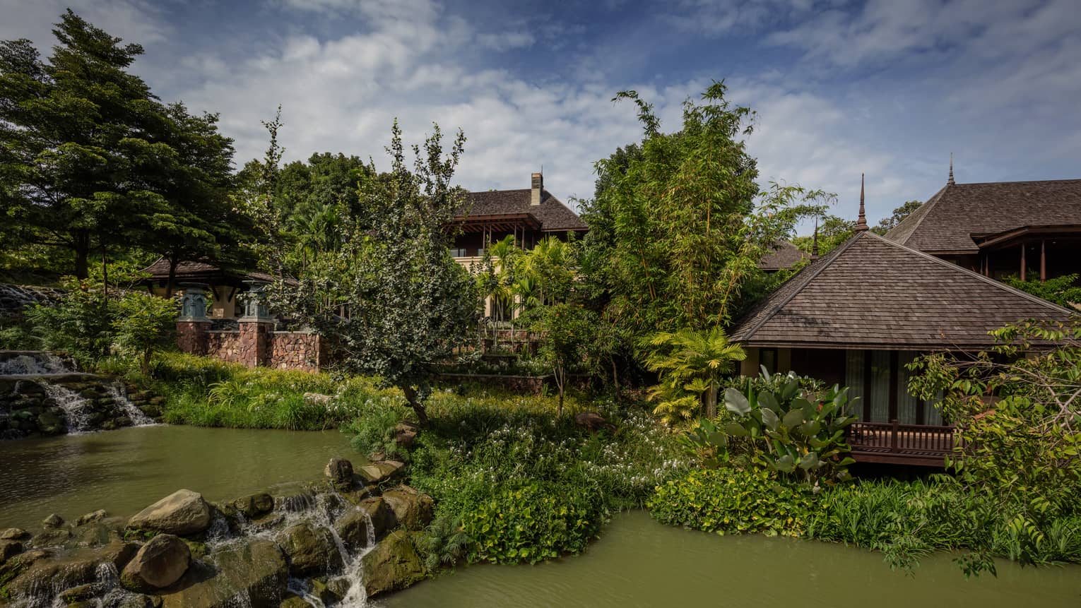 Tranquil resort villas surrounded by lush greenery waterfalls and a peaceful pond under a partly cloudy sky