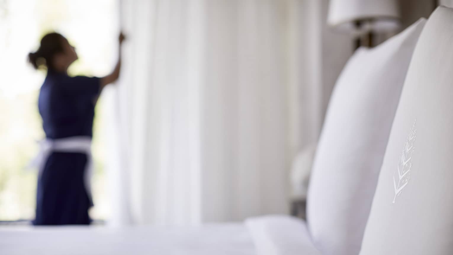 Close-up of bed, pillow with silhouette of hotel staff in uniform closing white curtains in background
