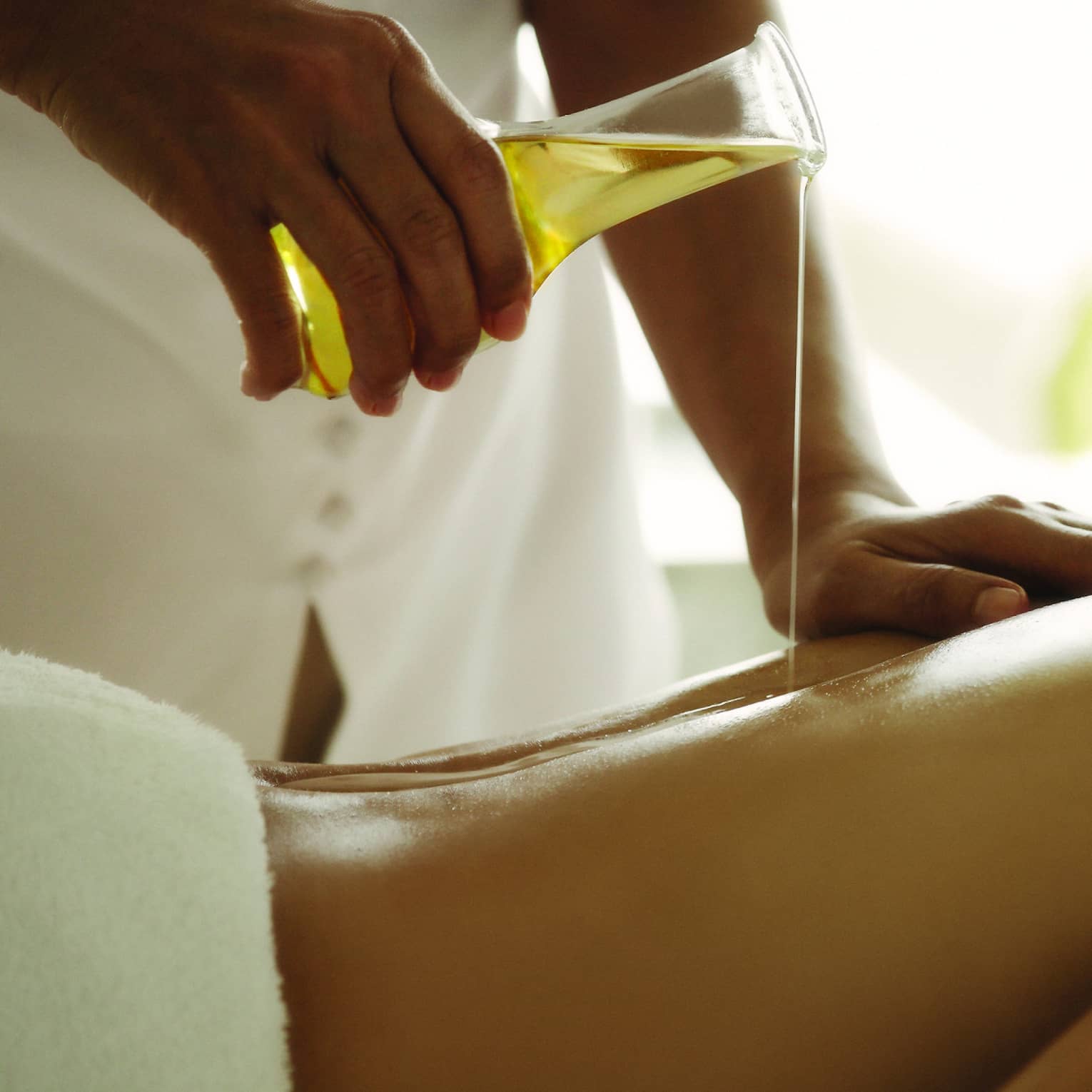 Spa staff pours massage oil over woman's bare back as she lays on treatment bed