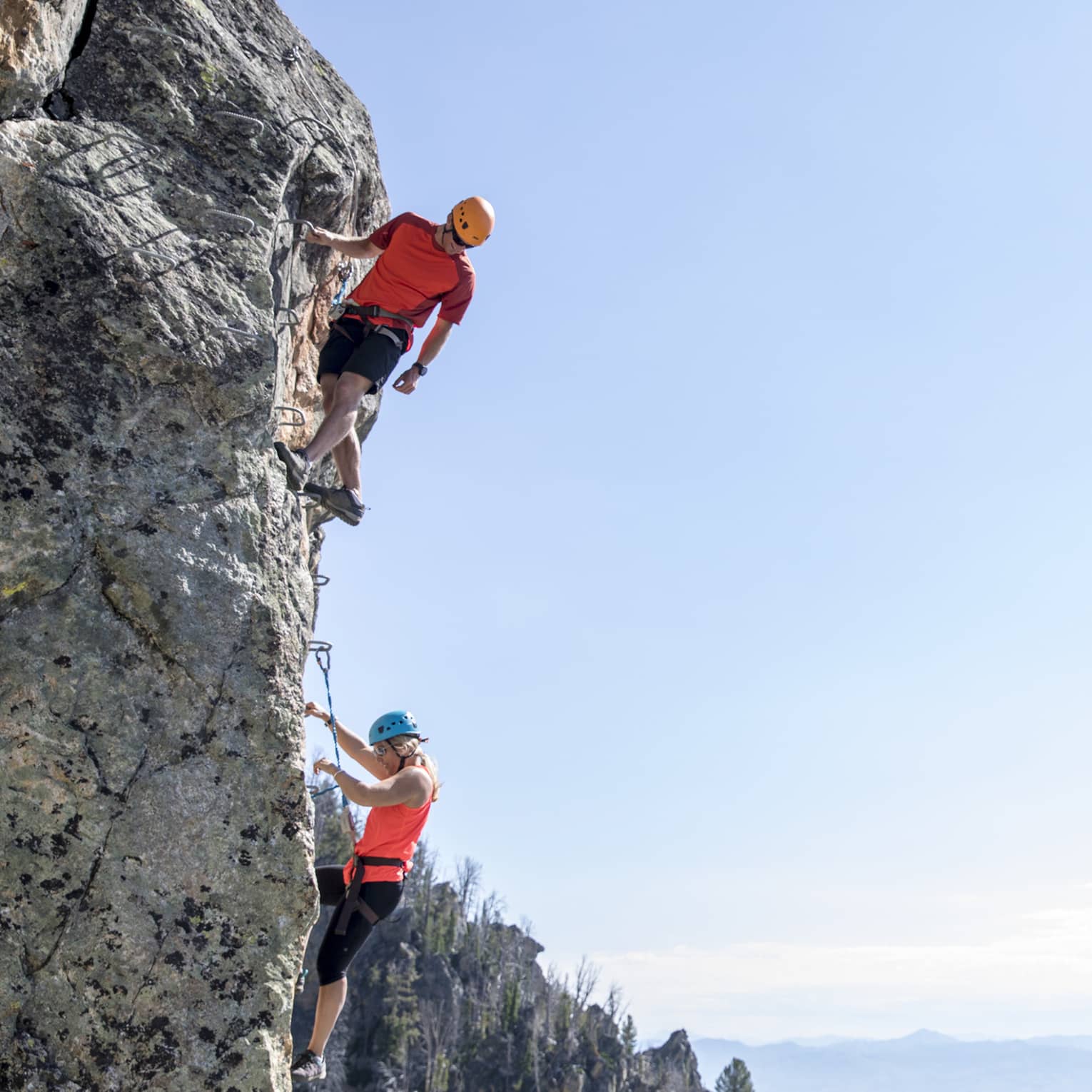 Man and woman climb steep cliff face with view of mountains in far distance 