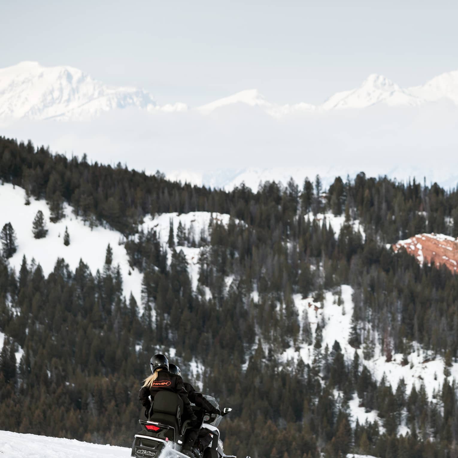 Couple rides snowmobile over mountain slope, trees and mountains in background