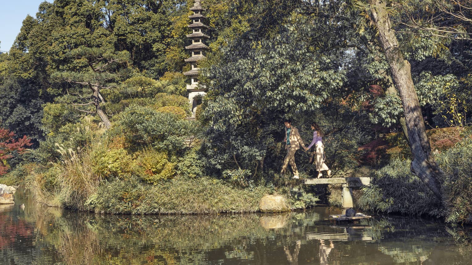 A couple walks on a bridge through a garden landscape by a pond