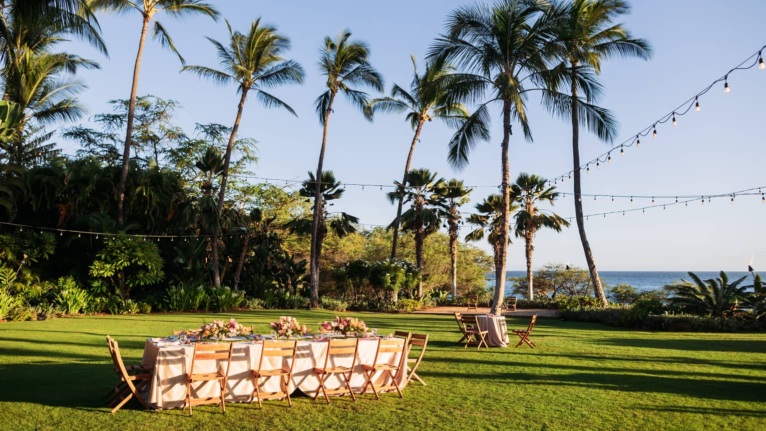 An outdoor banquet table in a large oceanfront green space decorated with colorful floral arrangements and string lights