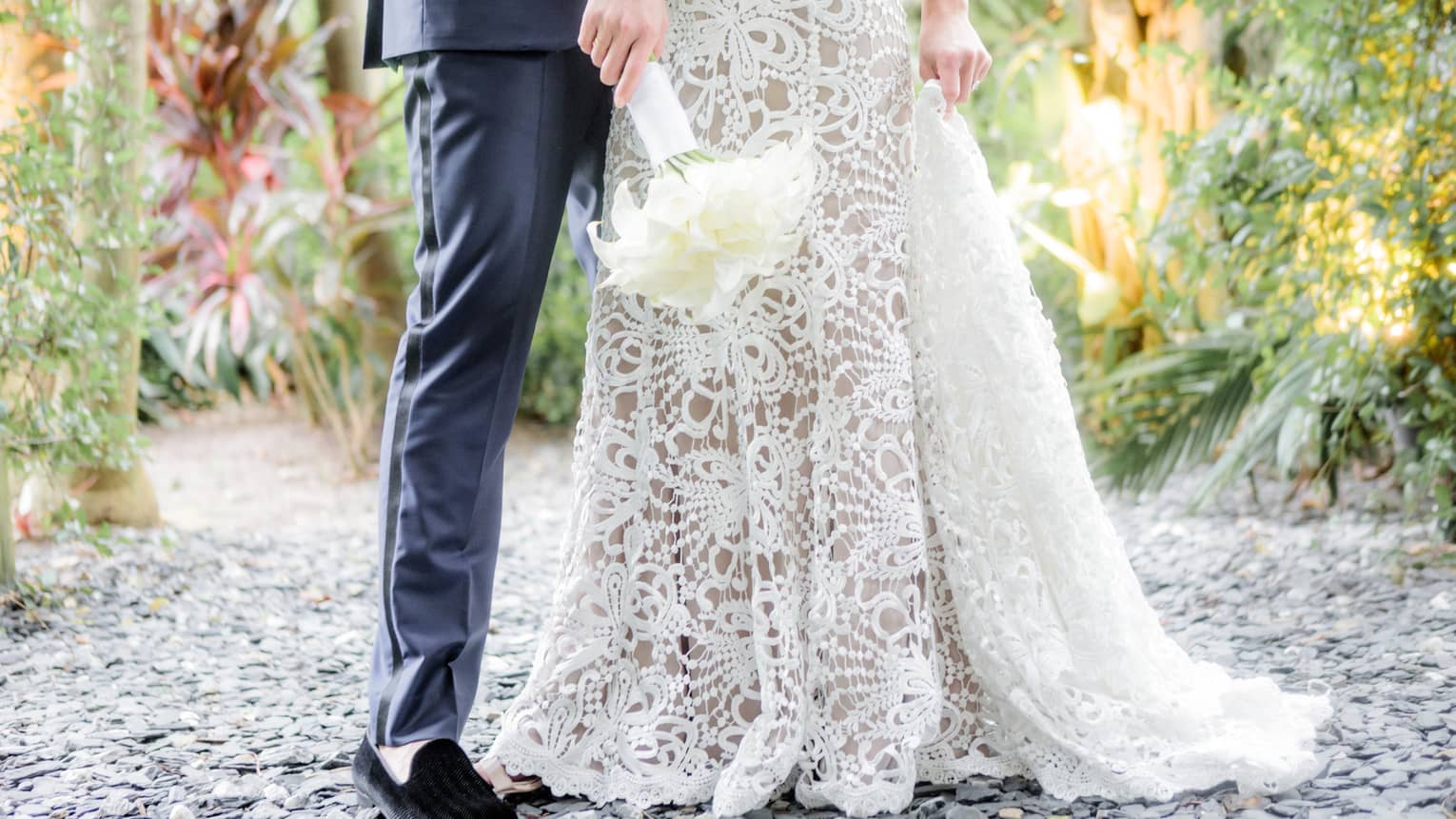 Lower view of bride's wedding gown skirt, groom's pants and shoes in tropical garden