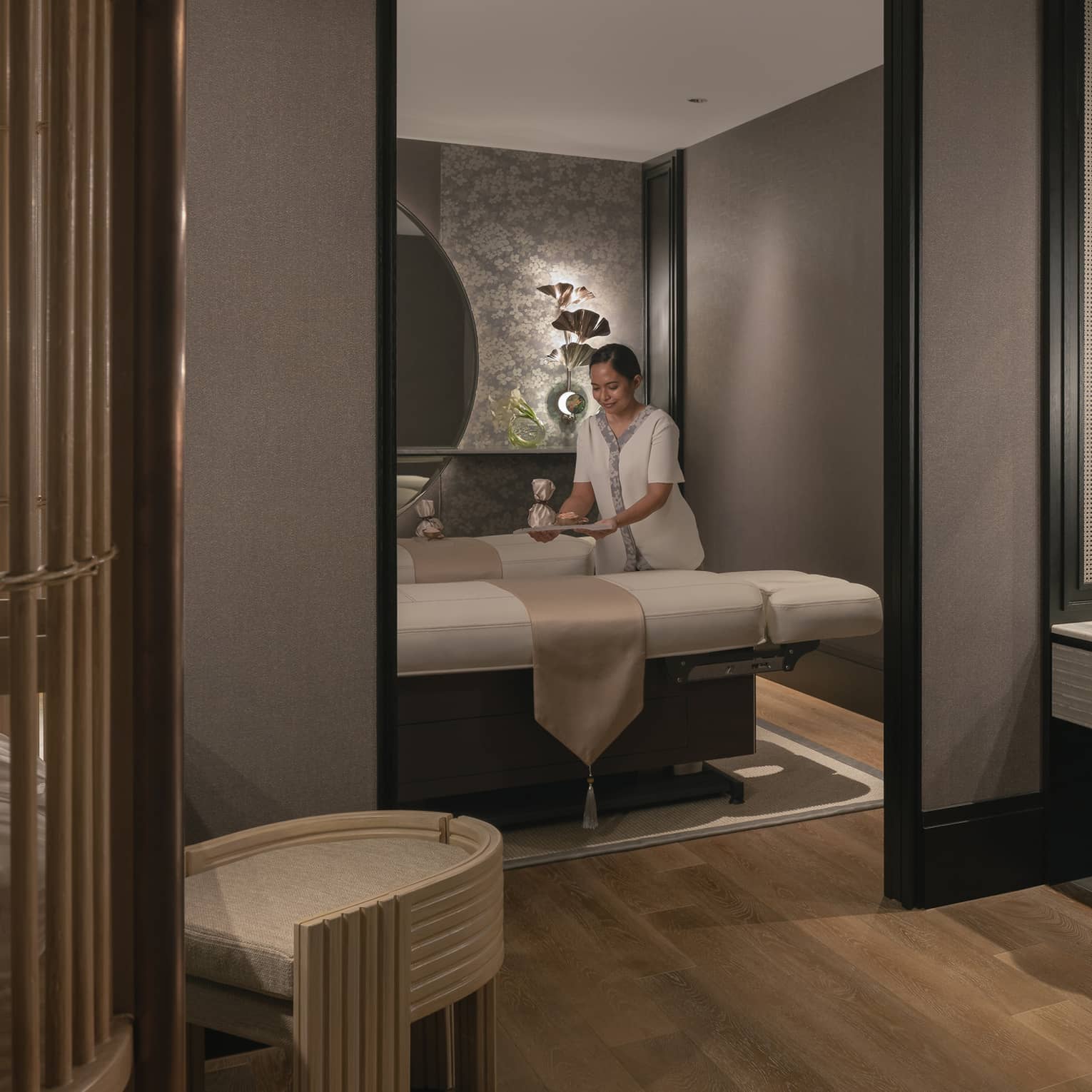 A tray is gently placed on one of two massage tables in a warmly lit spa room with soft grey walls and natural wood décor
