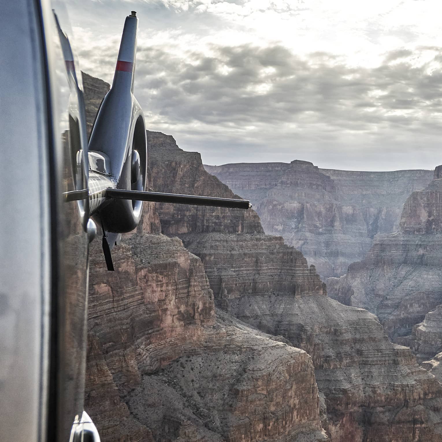 Viewed from a helicopter in flight, an awe-inspiring canyon landscape stretches into the distance beneath a cloudy sky.