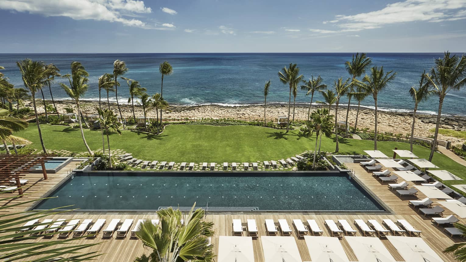 Aerial view of large outdoor swimming pool lined with white deck chairs, lawn, beach