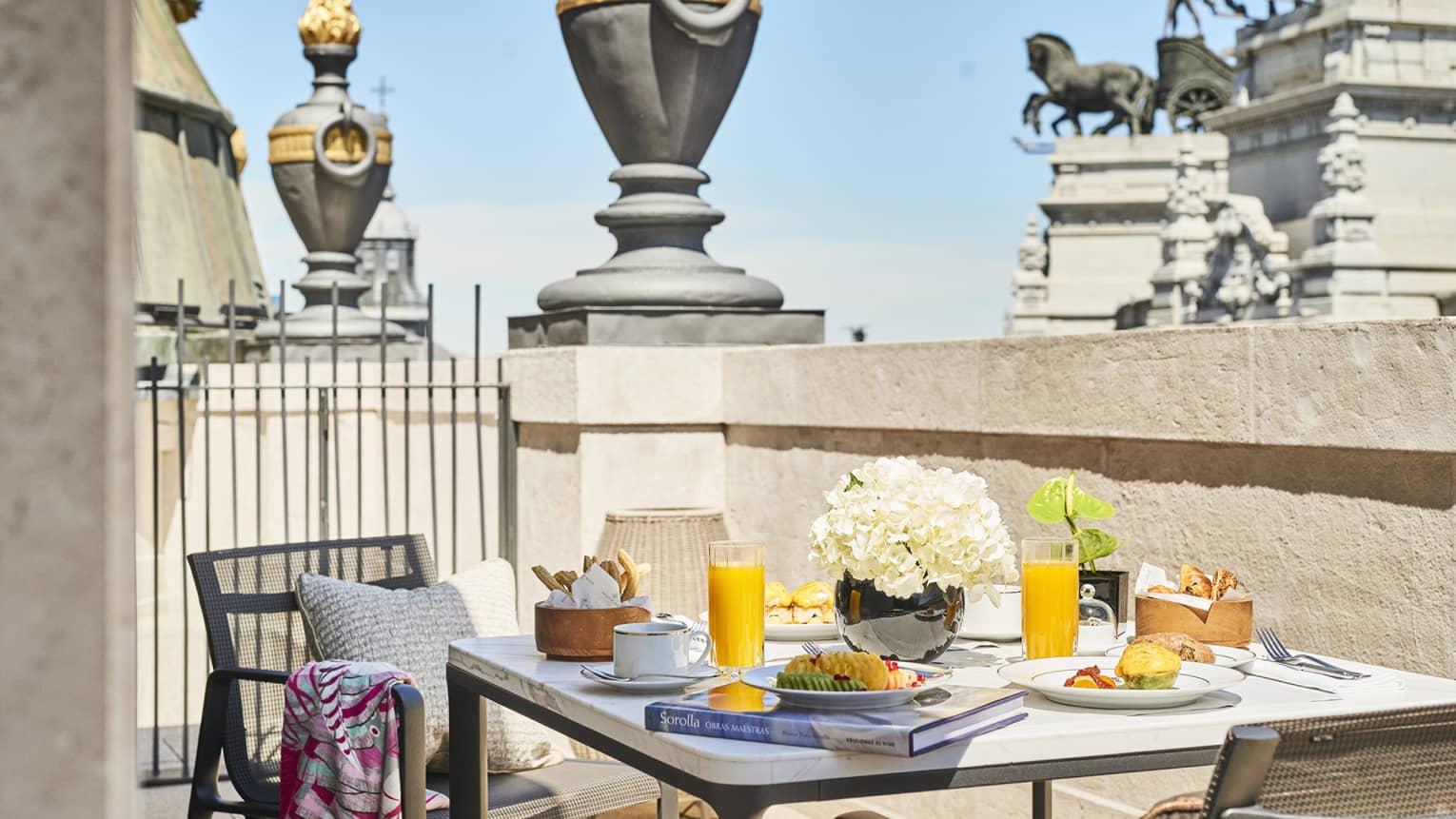 Breakfast setting on a luxury hotel terrace in Madrid with a table featuring orange juice, pastries, flowers and a book, overlooking historic architecture 