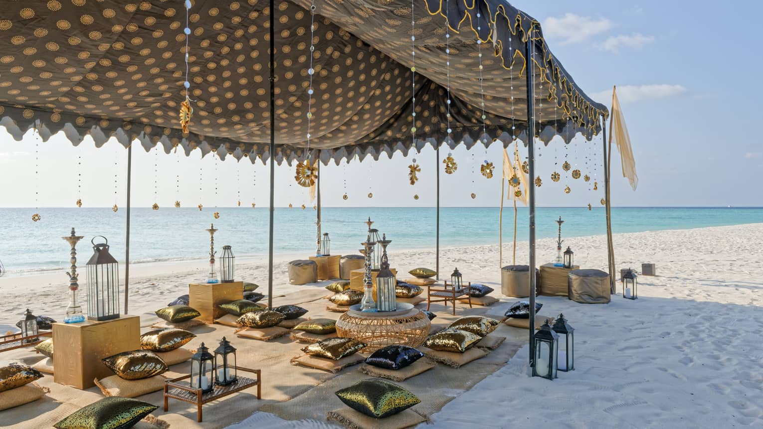 A tented shish-a bar set up on the beach with blankets and cushions.