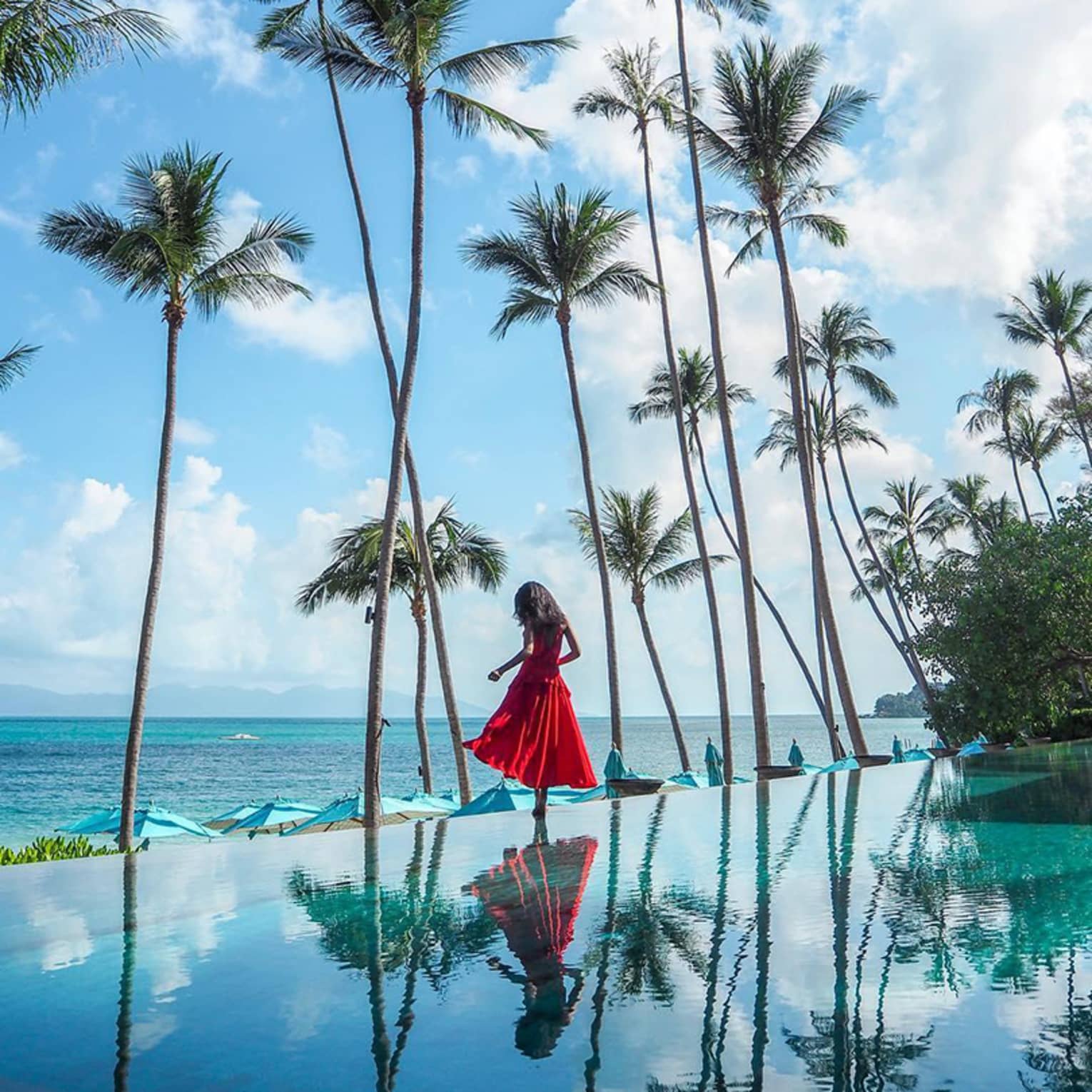 Girl wearing red dress stands at edge of infinity swimming pool under palm trees