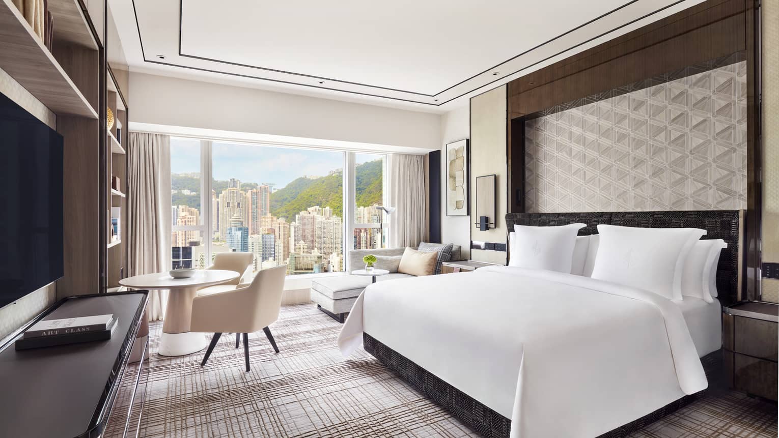 Modern hotel room with a large bed, geometric wall design, and a seating area next to a round table. Floor-to-ceiling windows offer a view of skyscrapers.