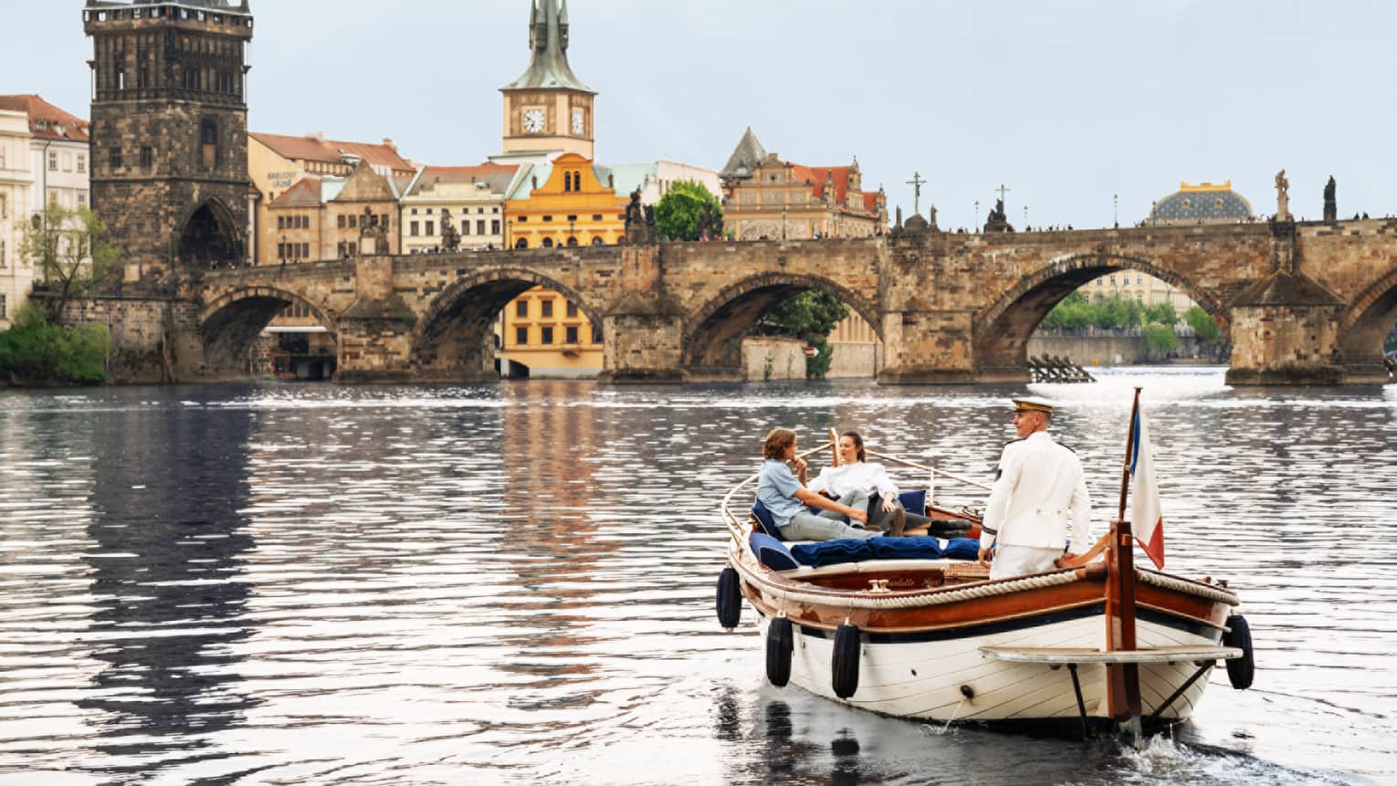 Couple with captain in wooden boat on Vltava River with bridge and buildings in backdrop