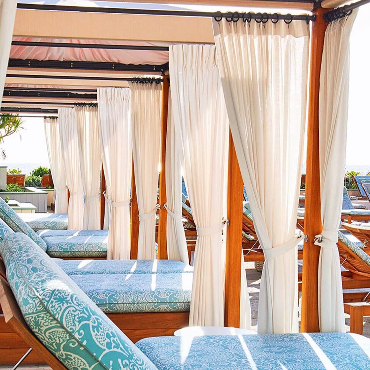 A row of patio lounge chairs with plush blue cushions under cabanas with white curtains, creating a luxurious and relaxing outdoor space.