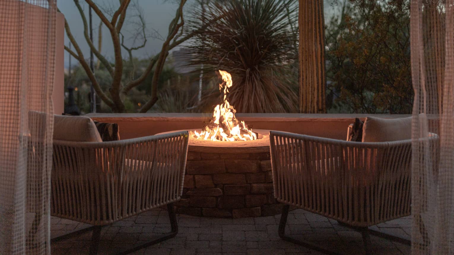 Two empty patio chairs in front of a lit fire pit in the evening