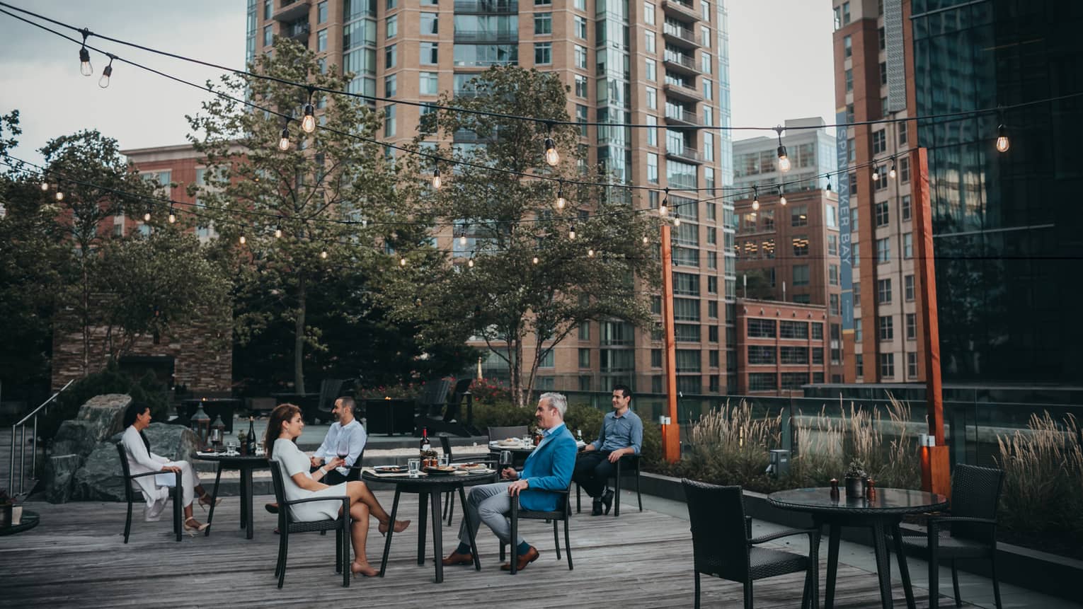 Three tables outside with a man and woman sitting at each enjoying a meal in the evening, there are trees and tall buildings in the background.