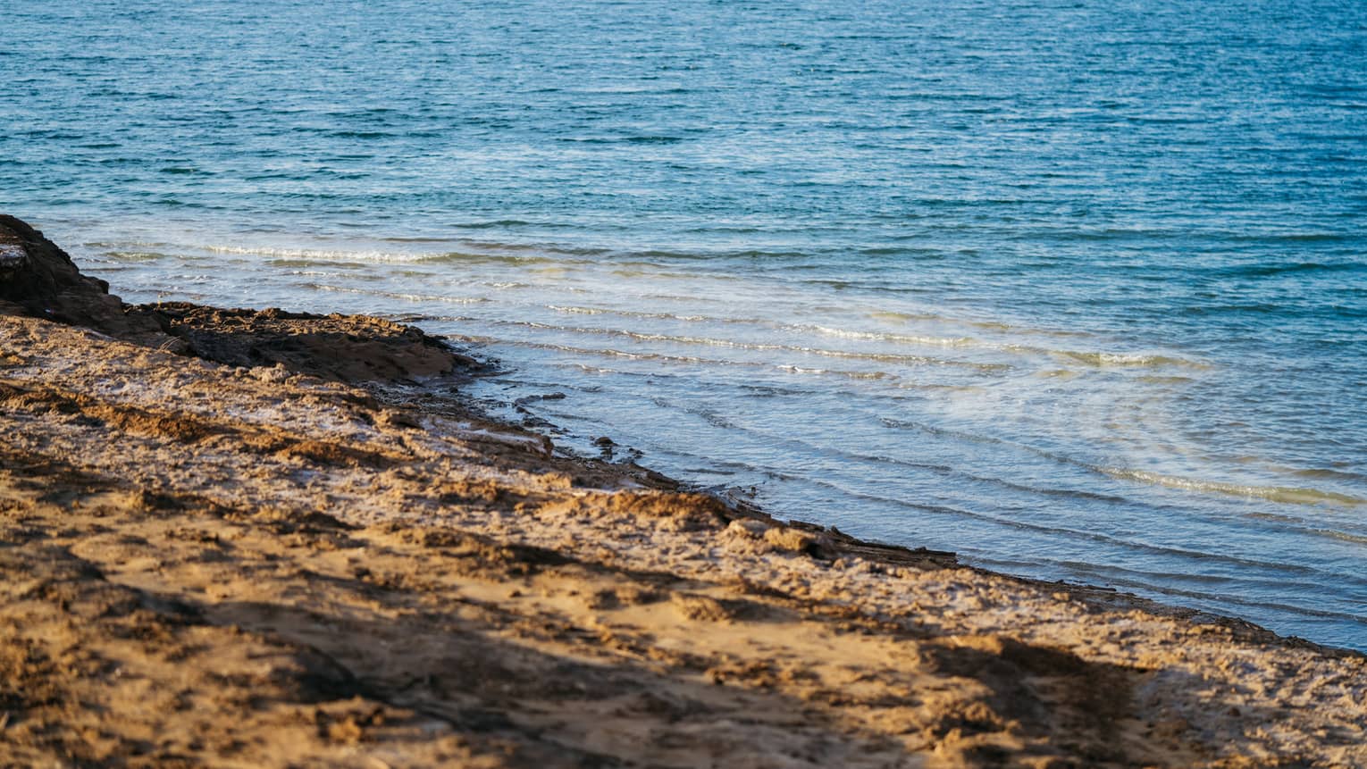 Gentle waves in sparkling azure water undulate toward an empty dark-brown, rough-sand beach.