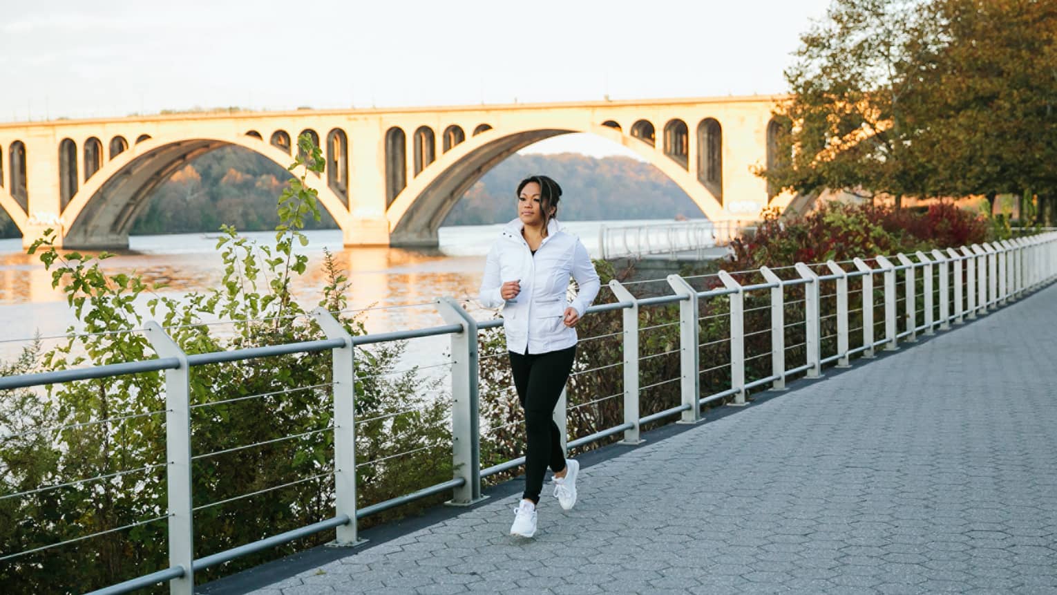 A woman in a white jacket jogging outside with a large bridge behind her.