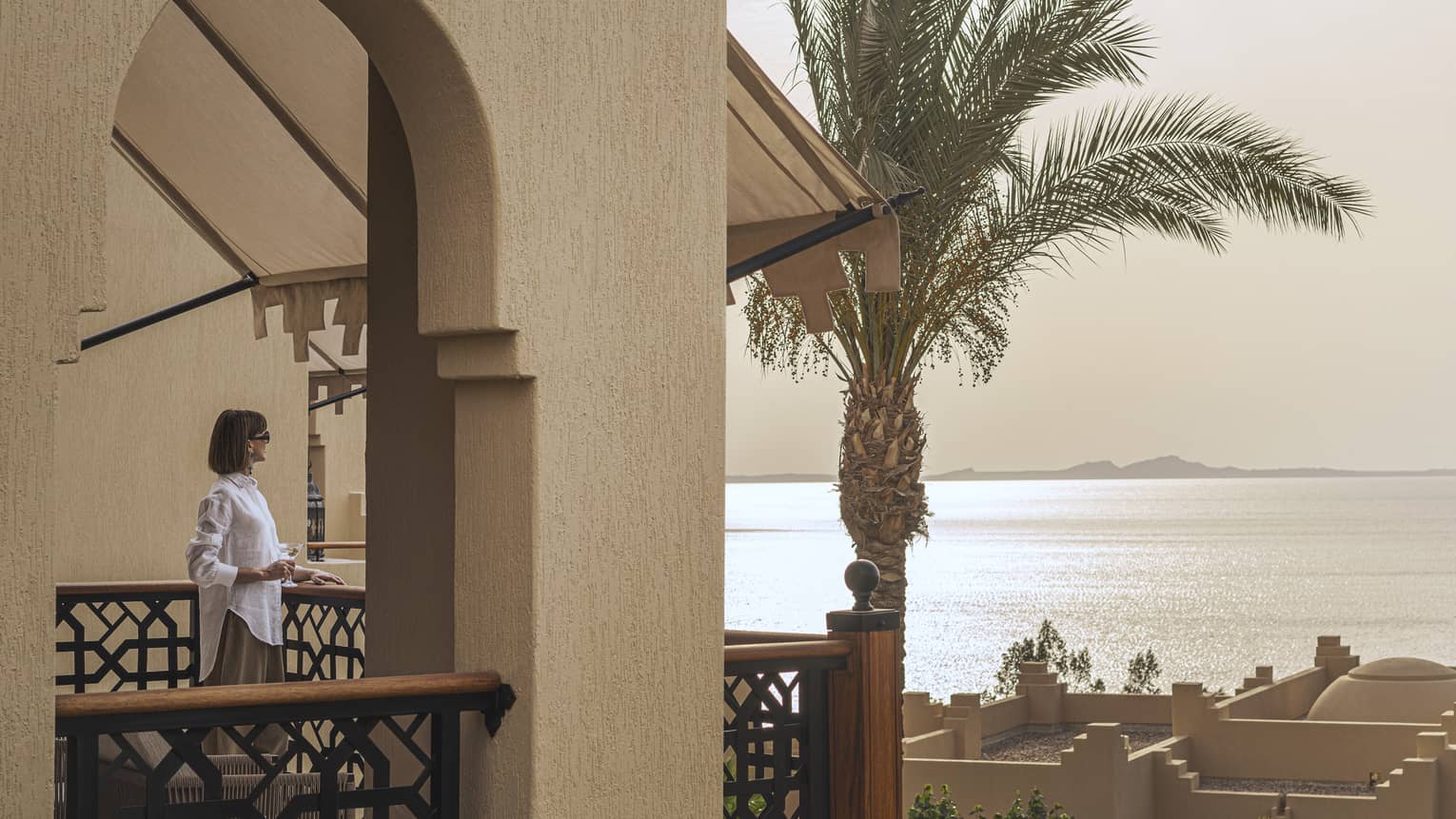 Woman in white shirt and sunglasses standing on room?s balcony gazes out at the Red Sea at sunset