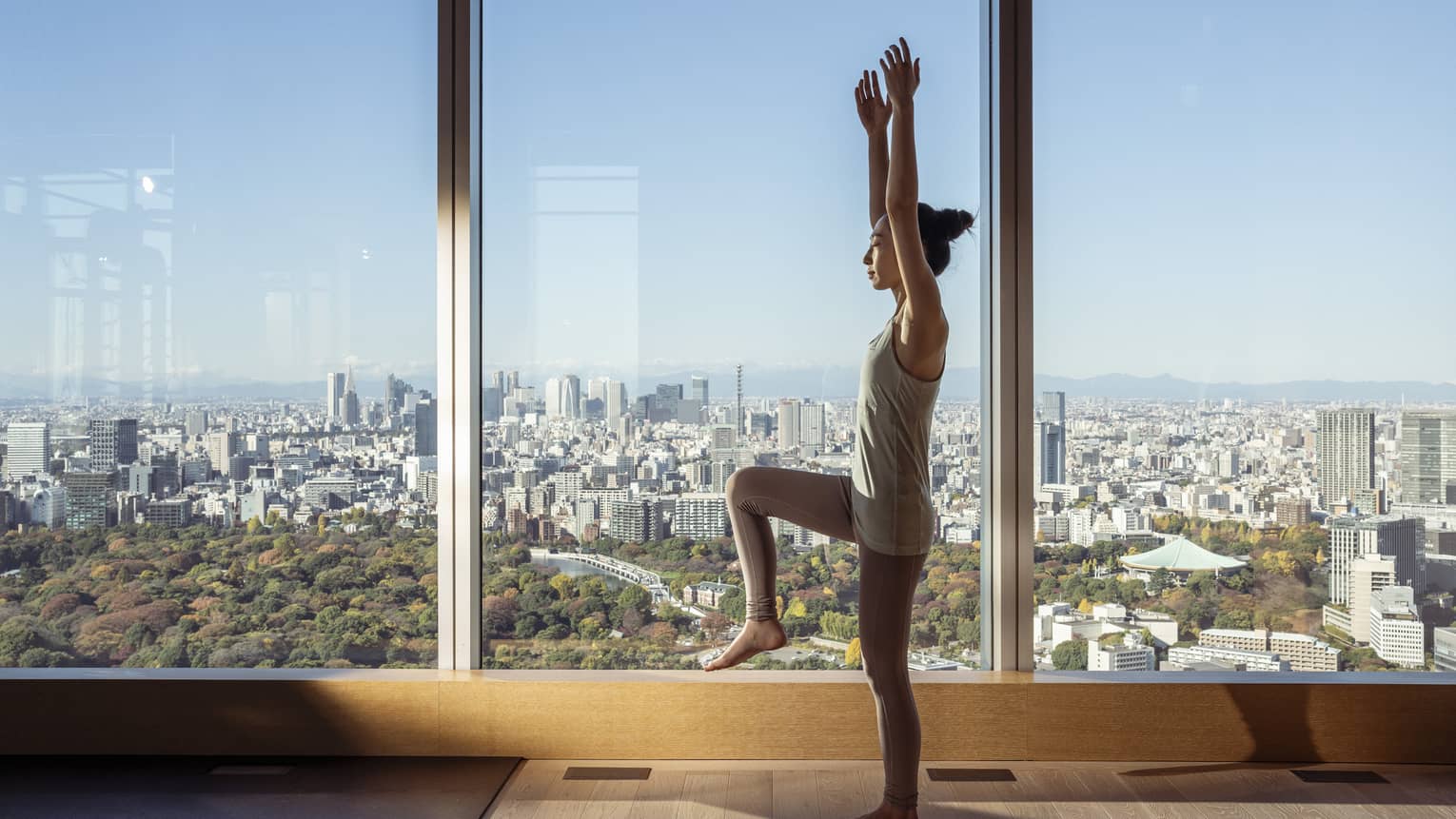 Woman stands in yoga pose in room with floor-to-ceiling windows with city views